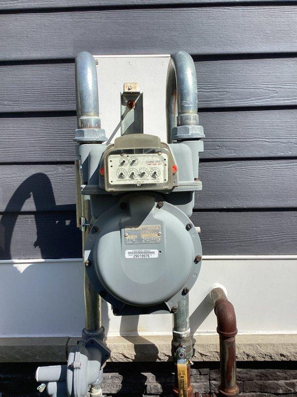 Conducted site assessment for whole house generator installation to replace customer's existing portable generator unit. Evaluated existing electrical service panel, gas meter assembly, and utility infrastructure to determine optimal placement and connection requirements for an 11kW single-phase automatic standby generator system. Documented current gas metering equipment, electrical meter location, and available space on exterior wall with dark gray siding. Identified adequate gas supply line accessibility and confirmed electrical service compatibility for generator integration. Photographed existing utility connections including gas regulator assembly, electrical meter box with PVC conduit, and surrounding landscape features to assess clearance requirements and installation constraints. Prepared comprehensive estimate for generator procurement, transfer switch installation, gas line extension, electrical panel modifications, and automatic transfer system integration to provide seamle