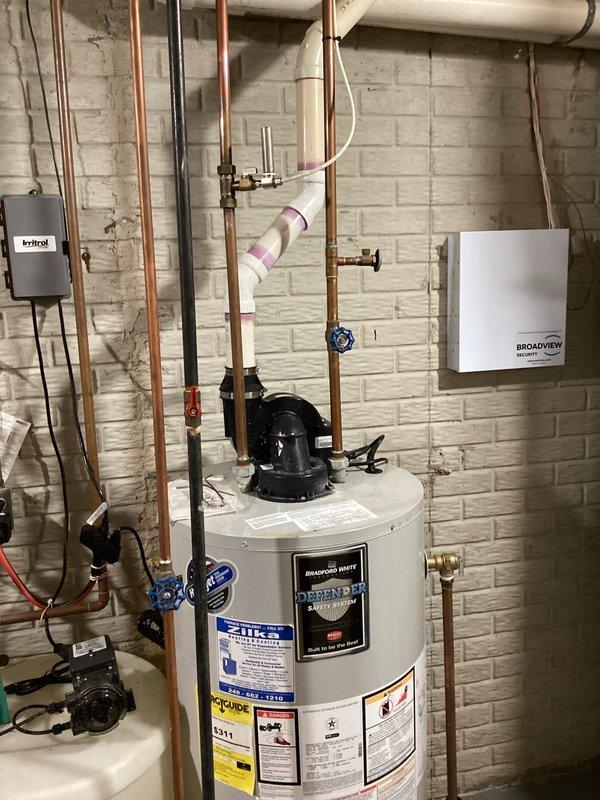 Completed comprehensive water heater assessment in basement utility area. Documented existing Bradford White water heater installation with associated copper supply lines, shut-off valves with blue handles, gas connections, and pressure relief components. Evaluated current plumbing configuration including vertical copper piping, PVC drainage system, and connection points for estimate preparation.