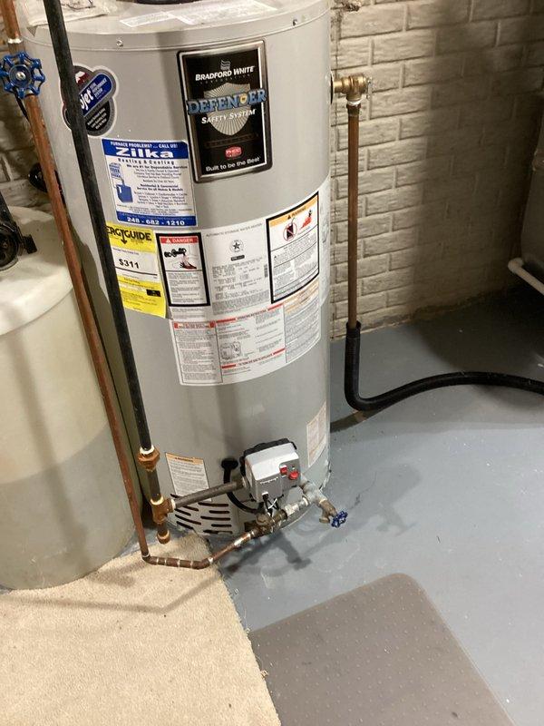 Completed comprehensive water heater assessment in basement utility area. Documented existing Bradford White water heater installation with associated copper supply lines, shut-off valves with blue handles, gas connections, and pressure relief components. Evaluated current plumbing configuration including vertical copper piping, PVC drainage system, and connection points for estimate preparation.