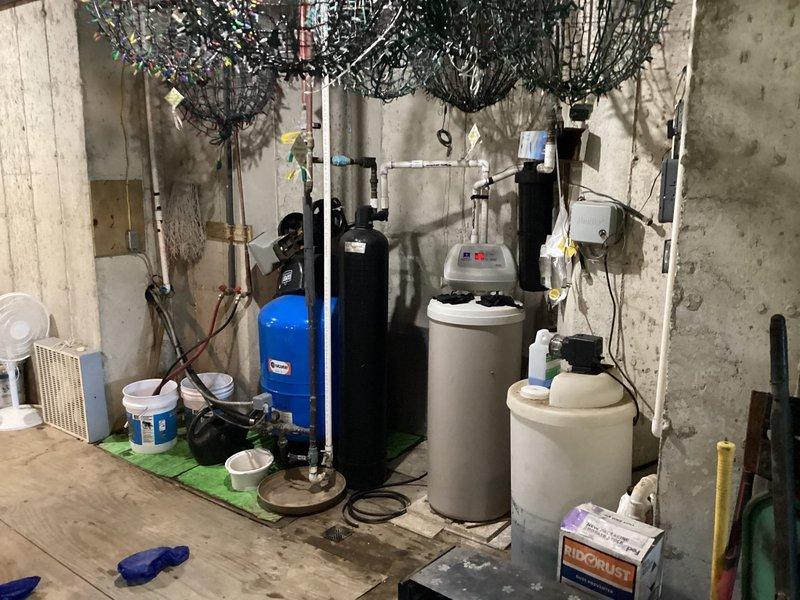 Responded to callback regarding oil contamination concerns on driveway and reports of oily water. Upon inspection, photographed basement mechanical room showing water treatment system including blue pressure tank, black filtration units, and white water softener tanks with digital control heads, all in operational condition with no visible oil leaks from HVAC equipment. Exterior inspection revealed concrete driveway with normal weathering and staining but no active oil discharge; no oil contamination detected on pavement or requiring cleanup at time of visit.
