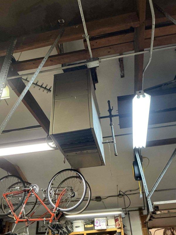 Inspected existing ceiling-mounted HVAC duct system in garage/workshop area. Evaluated rectangular metal air handler attachment points and support infrastructure. Identified potential upgrade opportunity where large cylindrical galvanized duct penetrates ceiling, noting suboptimal sealing and mounting hardware configuration. Recommended replacement of current ductwork with properly sized system including improved ceiling penetration seals and reinforced mounting brackets.