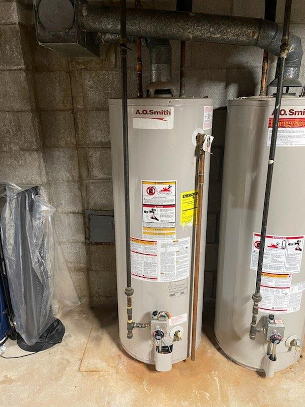 Conducted inspection of dual A.O. Smith water heater system in basement utility area. Units showed signs of aging including oxidation on copper pipes and mineral buildup on components. Evaluated flue pipe connection to ceramic base and checked integrity of red valve handle on left pipe assembly. System appears functional but exhibits normal wear consistent with regular use.
