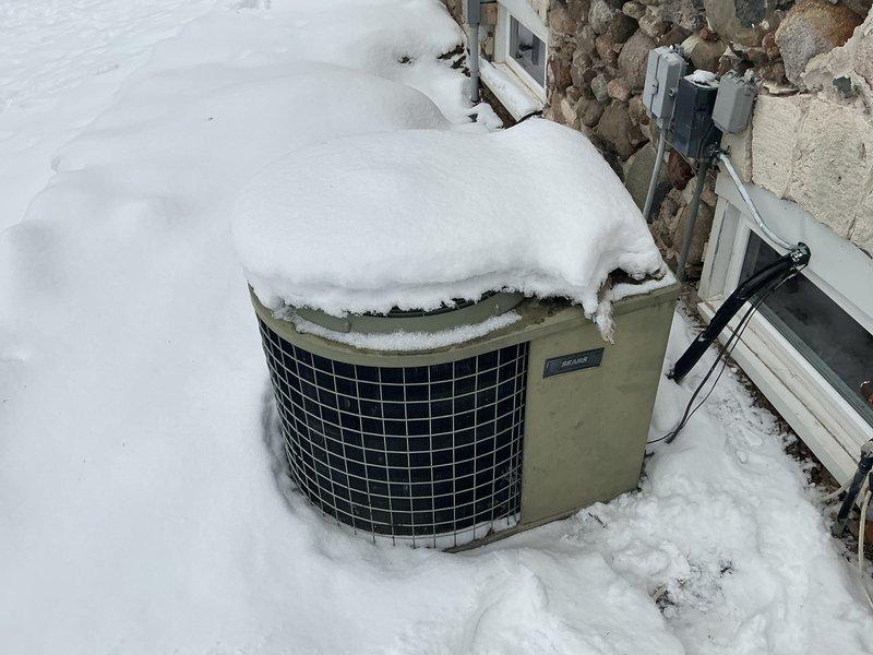 Conducted evaluation of a residential HVAC system requiring replacement due to multiple issues. The outdoor heat pump/AC unit was found severely compromised by excessive snow accumulation blocking proper airflow, potentially causing operating inefficiencies and possible damage. Indoor inspection revealed an aging furnace/air handler installed in an unfinished basement area with suboptimal installation conditions including proximity to bare stonework and unfinished flooring.