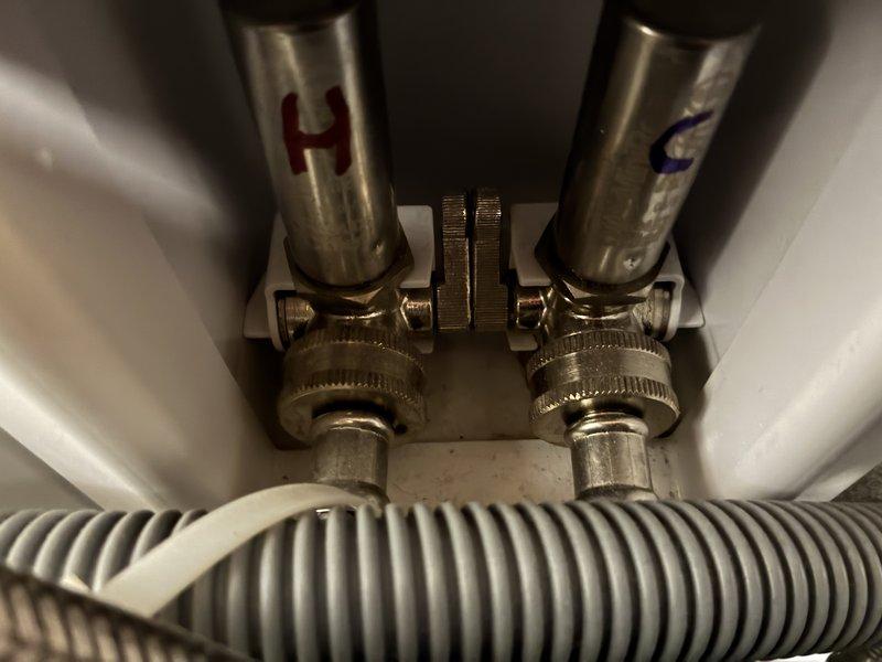 Inspected plumbing valves and connections in a mechanical system. Documented hot and cold water supply lines with proper labeling ("H" and "C" markings). Observed brass valve fittings and corrugated supply hose, noting some tarnishing on metal components. Visual inspection confirmed all components were properly installed and functioning as intended.