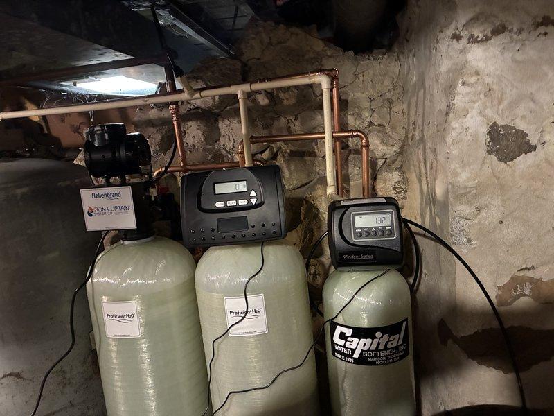 Completed installation of comprehensive water treatment system consisting of ProfessionalH2O water softener units and a Capital SOFTENER tank in basement utility area. System includes Solution Series electronic control panels with digital displays for monitoring water conditions. Connected all components to existing copper supply lines and verified proper function of all control units, including Hellenbrand pump components. Ensured integration with existing residential water heater and pressure tank for complete household water management solution.