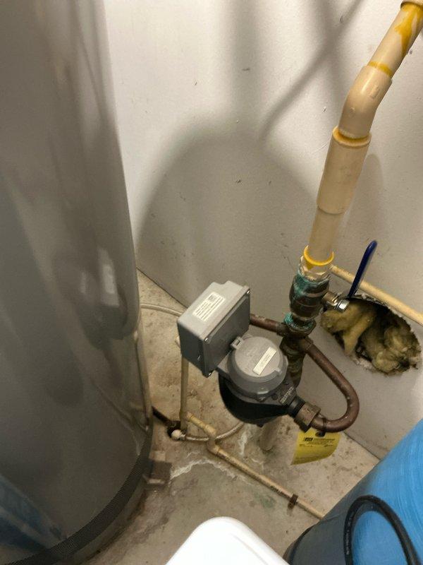 Completed HVAC system upgrade featuring installation of a new Aprilaire whole-home humidifier unit connected to existing ductwork. System includes copper piping with brass fittings and integrated digital thermostat control panel for improved climate regulation. Noted water damage around plumbing area requiring additional attention; recommended repair of exposed wall insulation adjacent to water meter.