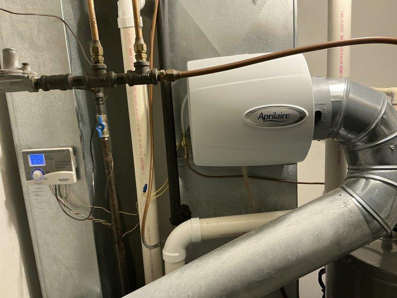 Completed HVAC system upgrade featuring installation of a new Aprilaire whole-home humidifier unit connected to existing ductwork. System includes copper piping with brass fittings and integrated digital thermostat control panel for improved climate regulation. Noted water damage around plumbing area requiring additional attention; recommended repair of exposed wall insulation adjacent to water meter.