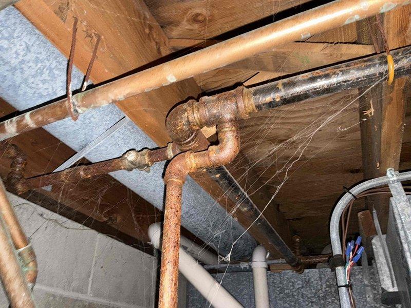 Conducted inspection of utility area revealing significant corrosion and oxidation on copper pipes in HVAC/plumbing system mounted beneath floor joists. Observed deterioration particularly severe at elbow joints, with visible rust compromising system integrity. Recommended complete replacement of affected copper piping and integration with existing PVC components to prevent potential leaks and system failure.