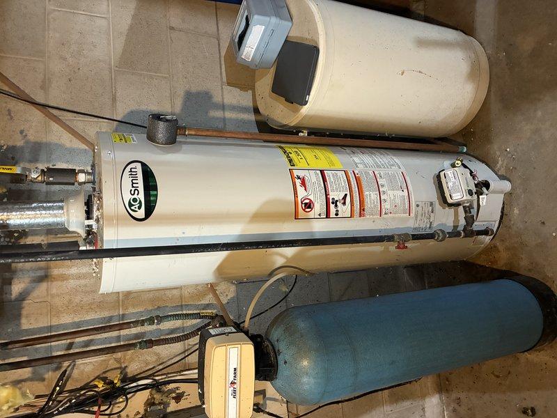 Inspected and documented A.O. Smith water heater installation with accompanying blue expansion tank in utility area. System features copper piping connections, standard electrical components, and proper safety labeling. Secondary filtration or water softening system observed adjacent to the main unit, all installed on tile flooring with appropriate wall-mounted electrical panel connections.