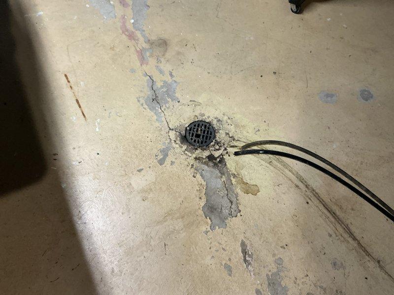 Conducted inspection of HVAC replacement site, documenting existing Amana furnace system in basement utility room with connected galvanized metal ductwork and ventilation PVC piping. Noted water damage issues around floor drain with visible staining, discoloration, and cracking that may impact installation area. Assessed current configuration for appropriate replacement specifications and ventilation requirements.