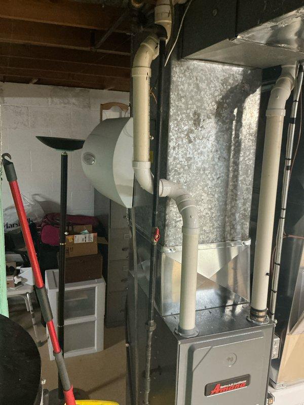 Conducted inspection of HVAC replacement site, documenting existing Amana furnace system in basement utility room with connected galvanized metal ductwork and ventilation PVC piping. Noted water damage issues around floor drain with visible staining, discoloration, and cracking that may impact installation area. Assessed current configuration for appropriate replacement specifications and ventilation requirements.