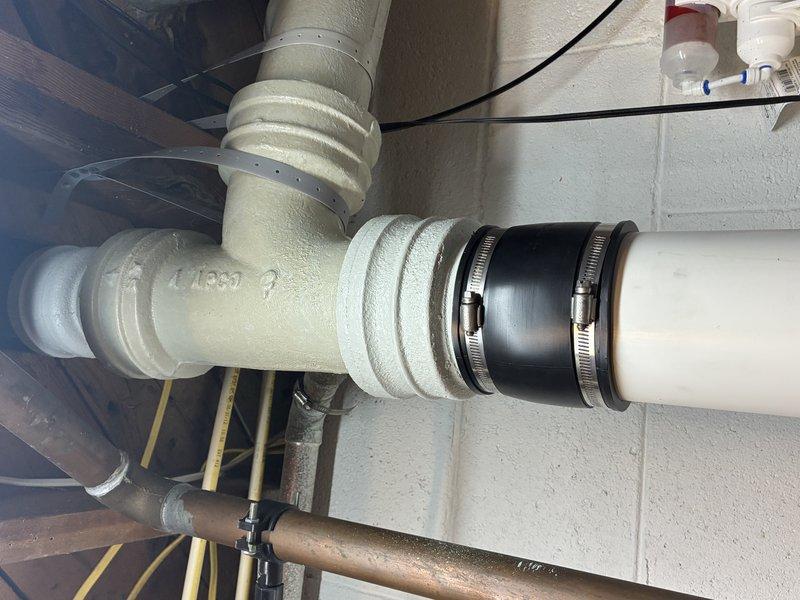 Conducted thorough assessment of basement plumbing infrastructure, documenting water supply system components including PVC piping configuration with multiple joints and fittings. Evaluated condition of T-junction pipe assembly and rubber coupling with metal band clamps securing vertical drainage sections. Verified integrity of all connection points and mounting hardware where pipes are secured to concrete wall structure.