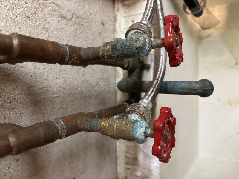 Conducted inspection of aging copper water supply lines exhibiting significant oxidation and corrosion. Identified brass fittings with tarnishing and examined red valve handles and flexible metal hoses for functionality. Evaluated black supply line with brass fitting and spiral metal protective covering, noting potential replacement requirements due to deterioration around connection points.