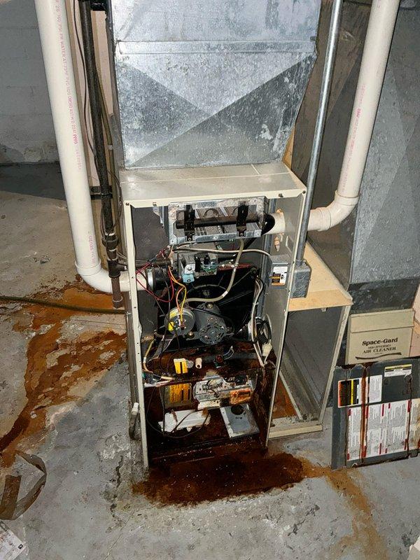 Inspected 15+ year old gas furnace displaying significant rust damage and leakage at the base, with extensive brown-red staining spread across the concrete floor indicating severe internal corrosion. System assessment revealed damaged heat exchanger and compromised components beyond economical repair. Recommended complete system replacement with new high-efficiency unit featuring upgraded filtration system to replace existing Space-Gard unit. Exterior inspection confirmed appropriate electrical service and venting options for replacement installation.
