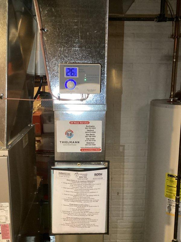 Successfully installed new high-efficiency furnace with digital thermostat display showing proper operation at 26 degrees. Mounted Aprilaire whole-home humidifier on the system, connecting all necessary tubing and ensuring proper integration with the HVAC system. Applied service identification sticker with 24-hour service contact information for future reference.