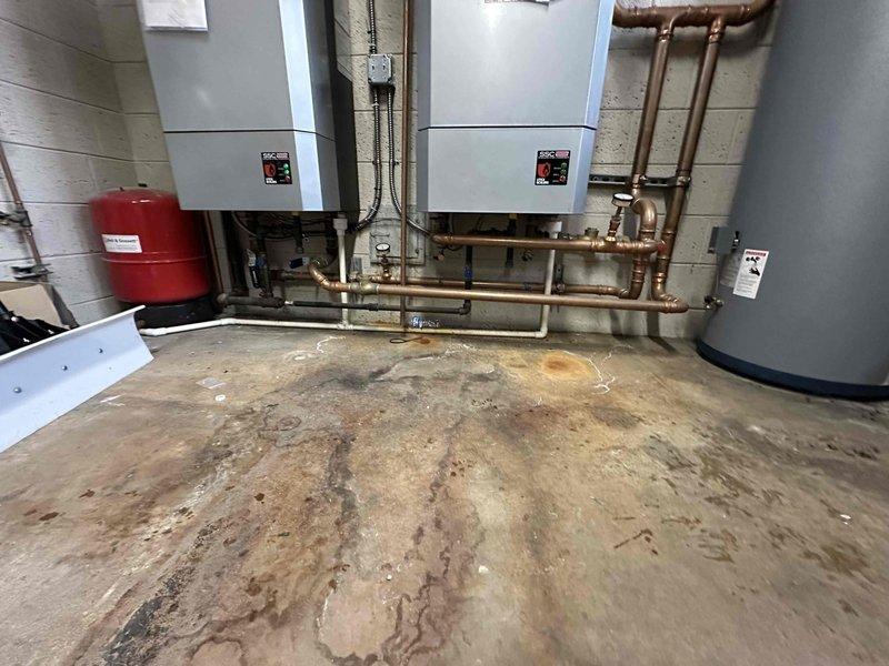 Conducted comprehensive assessment of existing dual wall-mounted boiler system with associated copper piping network and expansion tank. Documented significant water damage and mineral deposits on concrete floor, indicating potential previous leakage issues. Inspection revealed complex plumbing configuration with multiple control valves, pressure gauges, and insulated pipes connecting to cylindrical storage tank, requiring evaluation for system optimization or replacement.