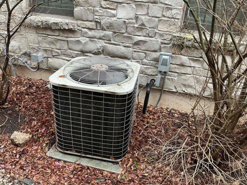 Completed assessment of an aging outdoor air conditioning condenser unit showing significant weathering and discoloration. Evaluated the installation site, which features limited accessibility due to narrow passageway between stone wall and wooden fence with accumulated debris and vegetation. Recommended full system replacement due to unit's condition and potential airflow restrictions in current location.