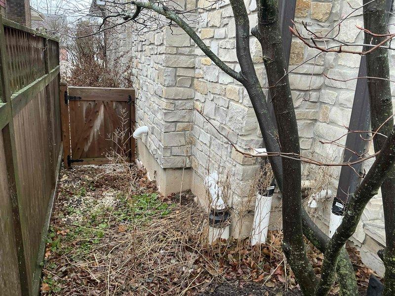 Completed assessment of an aging outdoor air conditioning condenser unit showing significant weathering and discoloration. Evaluated the installation site, which features limited accessibility due to narrow passageway between stone wall and wooden fence with accumulated debris and vegetation. Recommended full system replacement due to unit's condition and potential airflow restrictions in current location.