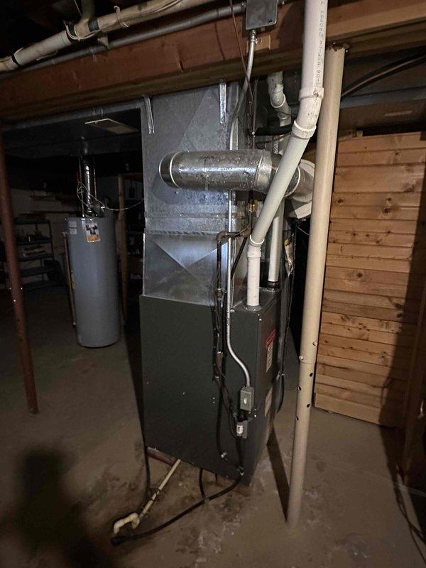 Inspected existing furnace installation in basement utility area. Documented current setup showing dark gray furnace with metal ductwork and PVC venting system. Evaluation focused on furnace condition, ventilation configuration, and surrounding connections including drainage components and copper piping for replacement estimation purposes.