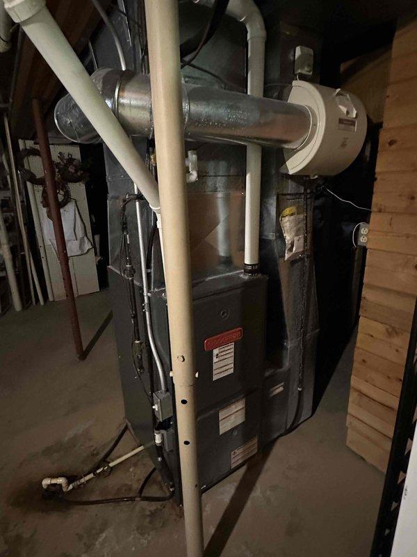 Inspected existing furnace installation in basement utility area. Documented current setup showing dark gray furnace with metal ductwork and PVC venting system. Evaluation focused on furnace condition, ventilation configuration, and surrounding connections including drainage components and copper piping for replacement estimation purposes.