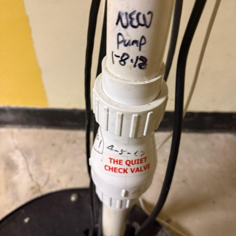 Inspected existing PVC plumbing system with check valve components. Observed previous installation work including "The Quiet Check Valve" and earlier pump replacement (noted as "NEW Pump 1-8-12"). Documented current condition of pipe connections, valve integrity, and minor mineral deposits on lower pipe sections. Photos taken of the installation for reference documentation.