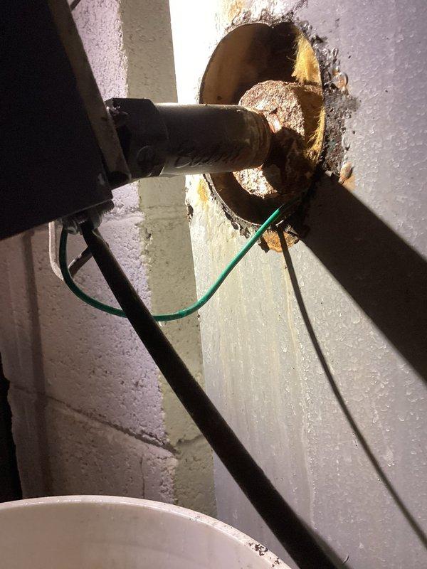 Identified and serviced severely corroded flange connection at plumbing penetration through cinder block wall. Observed significant rust deterioration around pipe entry point with compromised metal flange. Verified ground wire connection remained intact despite corrosion damage.