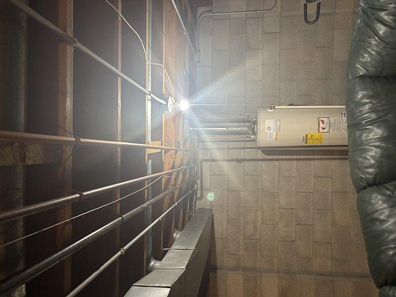 Conducted site assessment of water heater installation in utility room. Inspected existing plumbing infrastructure, including PVC/copper pipe runs along ceiling and wall framing. Evaluated water tank condition, connecting fixtures, and supporting brackets. All systems appear properly installed with appropriate insulation and safety labeling in place.