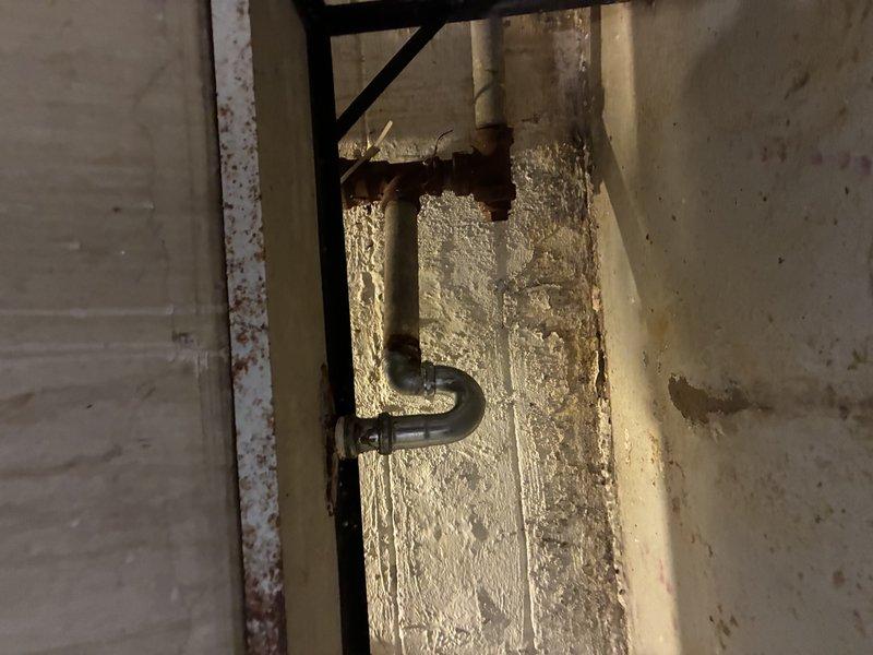 Conducted water service assessment of existing plumbing system. Inspected concrete basin containing water service connections showing significant mineral buildup and sediment accumulation. Evaluated drainage system including metal trap pipe and connected components for functionality; no active leaks detected despite aging infrastructure.