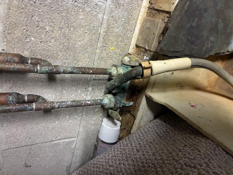 Performed inspection of residential water supply system, documenting significant copper pipe corrosion and water damage at connection points. Observed extensive oxidation with characteristic greenish-blue patina on copper pipes emerging from concrete floor, along with deterioration of surrounding materials and possible moisture-related damage. Recommended replacement of corroded supply lines and valve assemblies to prevent potential leaks and system failure.
