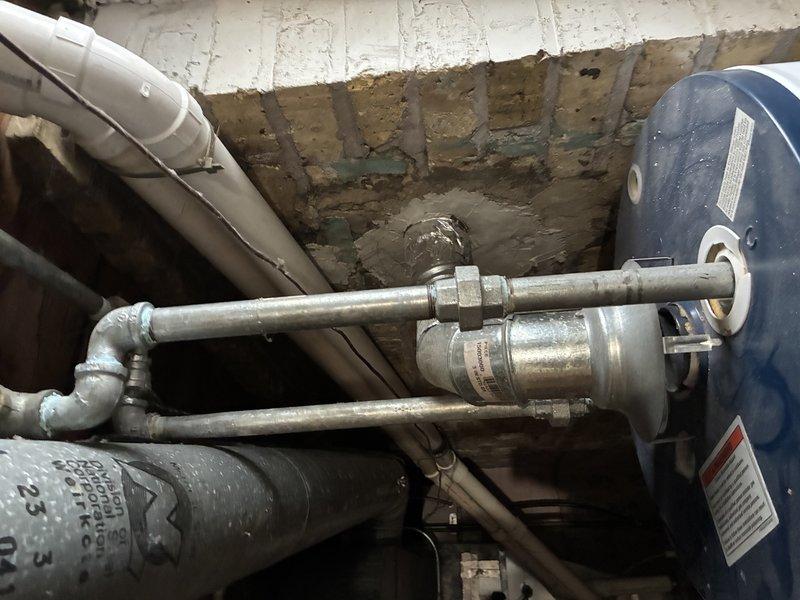 Conducted inspection of existing water heater system in basement utility area. Observed aging galvanized pipe connections to dark blue water heater tank with signs of surface corrosion at junction fittings. Noted potential issues with the cross-shaped junction and valve assembly. Provided estimate for replacement of deteriorating components and recommended upgrading to code-compliant PVC piping system for improved longevity and performance.