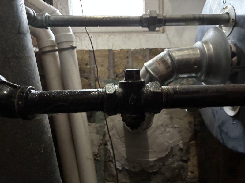 Conducted inspection of existing water heater system in basement utility area. Observed aging galvanized pipe connections to dark blue water heater tank with signs of surface corrosion at junction fittings. Noted potential issues with the cross-shaped junction and valve assembly. Provided estimate for replacement of deteriorating components and recommended upgrading to code-compliant PVC piping system for improved longevity and performance.