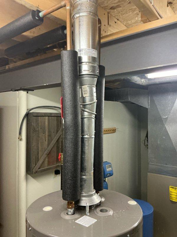 Completed assessment of water heater system in basement utility area. Observed PVC drainage pipe with visible mineral deposits on concrete floor and inspected vertical metal duct system with black foam insulation sleeves. Noted copper pipe branching horizontally from main assembly requiring attention.