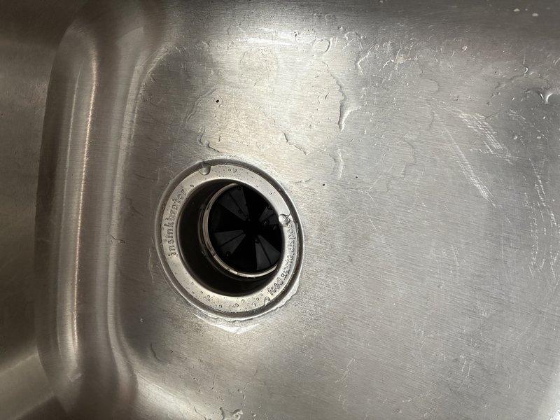 Inspected and documented customer's stainless steel sink with existing garbage disposal installation. System includes black disposal unit with proper rubber flaps in drain opening, connected to white PVC drain assembly with standard P-trap configuration. All components appear functional with normal wear patterns visible on sink surface.