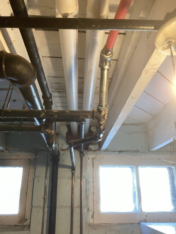 Conducted inspection of basement utility area to assess plumbing infrastructure connecting to HVAC system. Identified aging pipe joints with visible corrosion and mineral deposits at connection points between different pipe materials. Documented condition of support brackets and mounting hardware on ceiling installation for future reference.