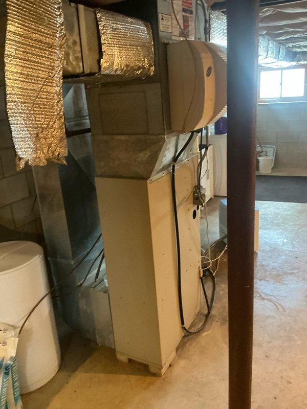 Conducted assessment of basement HVAC installation area featuring concrete flooring with existing drain access. Documented current air handler configuration with foil-faced insulated ductwork connected to vertical furnace unit. Evaluation completed for replacement estimate of aging system in utility room with concrete block walls.