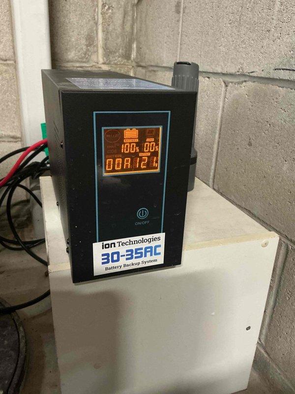 Installed and configured an Ion Technologies 30-35AC Battery Backup System for sump pump protection. System includes dual cooling fans, power supply, and indicator lights to ensure continuous operation during power outages. Mounted unit securely on wooden shelf against concrete block wall with proper electrical connections established.