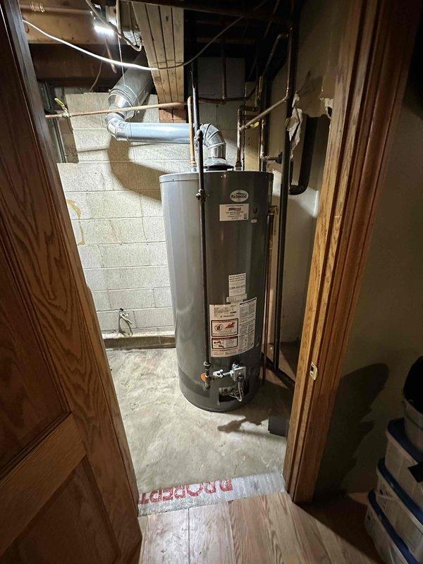 Conducted visual inspection of basement utility area containing water heater and associated ductwork. Evaluated existing cylindrical gray water heater installation, noting connection points to metal piping network and ventilation system. Assessed concrete block wall conditions and examined ceiling-mounted utility connections for potential replacement considerations.