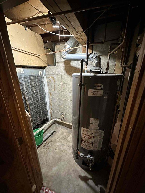 Conducted visual inspection of basement utility area containing water heater and associated ductwork. Evaluated existing cylindrical gray water heater installation, noting connection points to metal piping network and ventilation system. Assessed concrete block wall conditions and examined ceiling-mounted utility connections for potential replacement considerations.