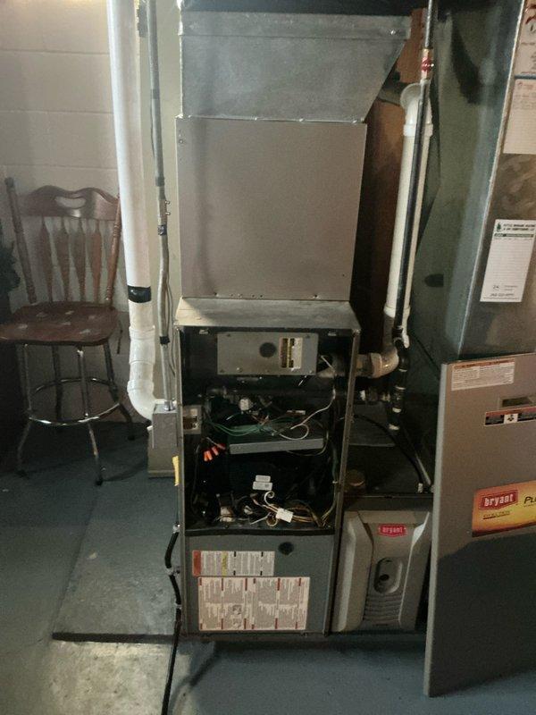 Inspected Bryant furnace unit with front panel removed for comprehensive system evaluation. Observed exposed control board, internal wiring, and connected ductwork. Noted electrical wiring issue requiring attention where single black wire protrudes through floor with visible rust/corrosion around penetration point.