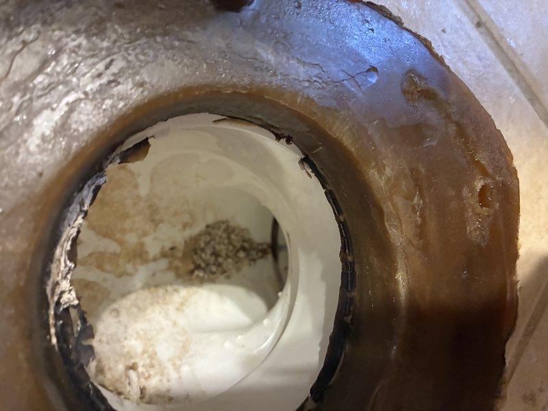 Inspected toilet flange showing significant deterioration with visible rust, corrosion and mineral buildup. Documented damaged white plastic flange with brownish residue and sediment accumulation around the central opening. Recommended replacement of the deteriorated flange to prevent potential leaks and ensure proper toilet mounting.