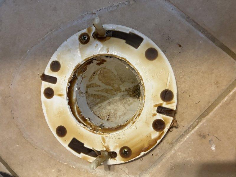 Inspected toilet flange showing significant deterioration with visible rust, corrosion and mineral buildup. Documented damaged white plastic flange with brownish residue and sediment accumulation around the central opening. Recommended replacement of the deteriorated flange to prevent potential leaks and ensure proper toilet mounting.