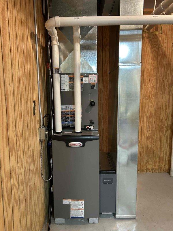 Completed installation of a new Lennox HVAC complete system in the utility area. Mounted main furnace unit on concrete floor against wood-paneled wall, connected proper PVC ventilation piping with multiple elbows extending upward. Installed and connected all required ductwork with appropriate metal fittings to ensure optimal system performance.