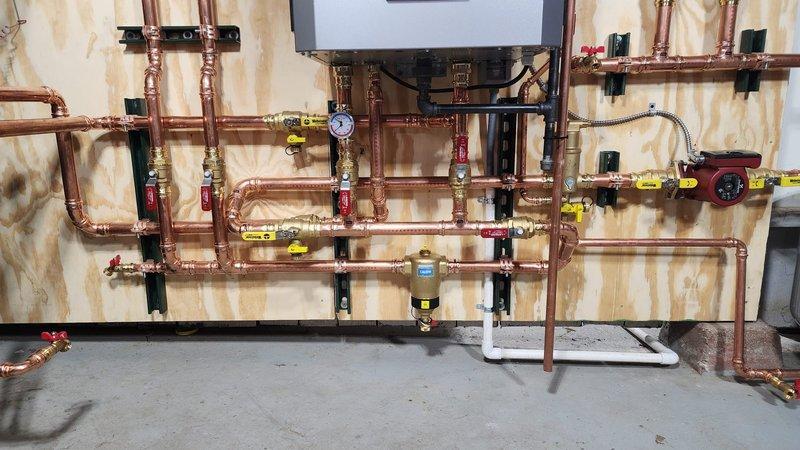 Completed installation of an NTI boiler system mounted on plywood backing with comprehensive copper pipe network. Installation included pressure gauge, multiple shut-off valves with identification tags, circulation pump, and digital control panel programming. All connections were properly secured with appropriate mounting brackets and system was tested for proper operation.
