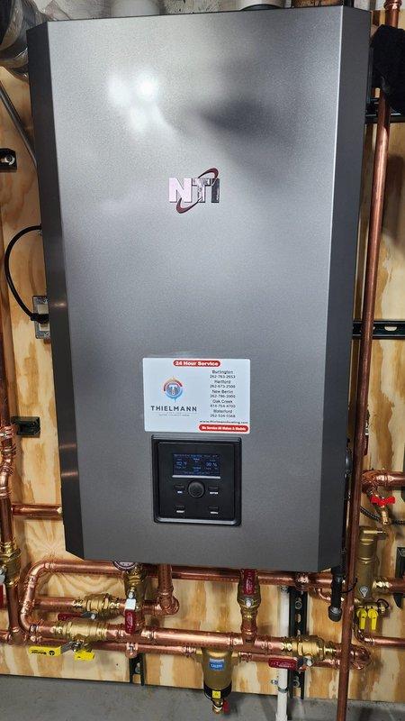 Completed installation of an NTI boiler system mounted on plywood backing with comprehensive copper pipe network. Installation included pressure gauge, multiple shut-off valves with identification tags, circulation pump, and digital control panel programming. All connections were properly secured with appropriate mounting brackets and system was tested for proper operation.