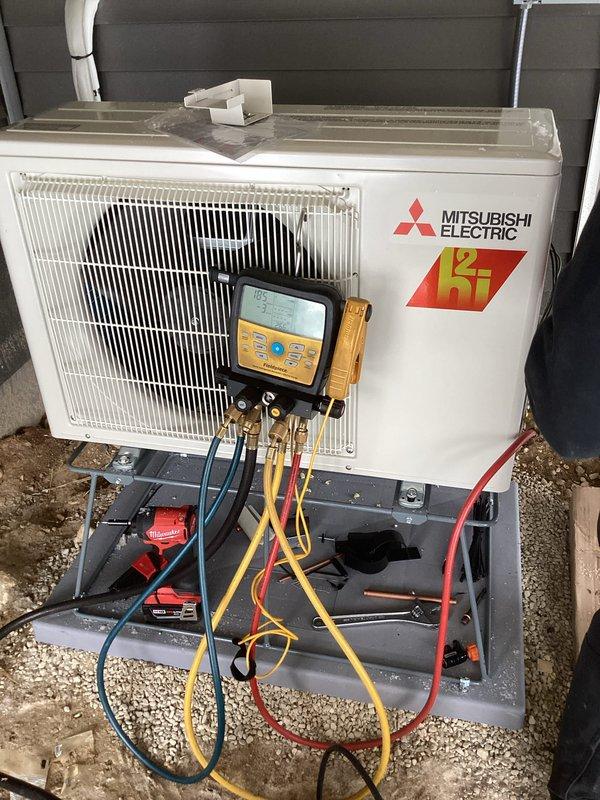 Completed installation of Mitsubishi Electric mini-split system with outdoor condenser unit properly set on concrete pad with gravel base. Indoor head unit was wall-mounted in construction space with exposed ceiling joists. System was pressure tested, evacuated, and charged according to manufacturer specifications with final diagnostic verification confirming proper operation.