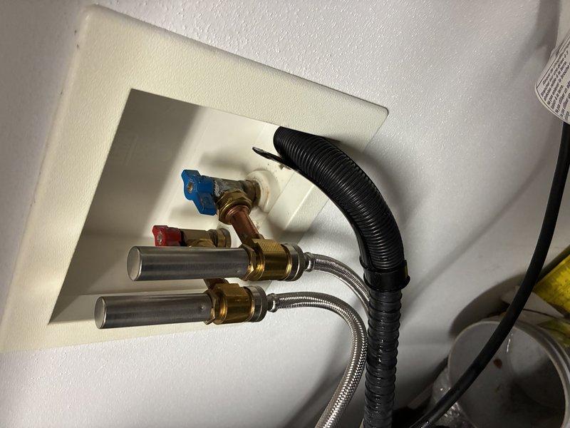 Installed and inspected water supply connections in recessed wall box, including two shut-off valves (hot and cold) with brass fittings and stainless steel braided supply lines. Verified proper installation of drainage system with black corrugated hose and confirmed no leaks or corrosion present in the entire assembly.