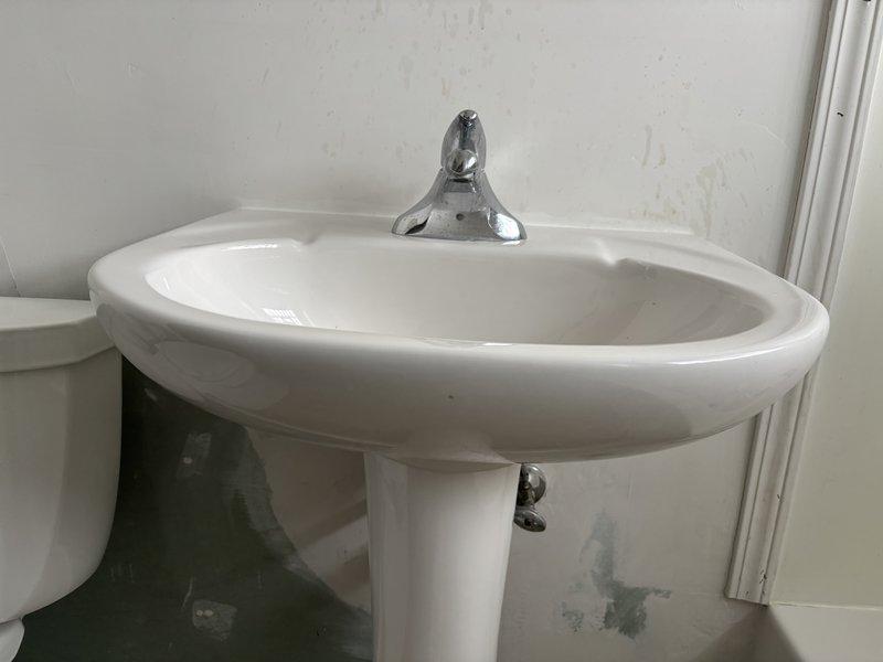 Inspected bathroom fixtures including pedestal sink and surrounding area. Identified minor water damage and discoloration where sink meets wall and floor. No significant plumbing issues with toilet visible in assessment photos, though partial view suggests standard installation and condition.