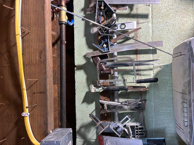 Inspected exposed plumbing infrastructure in utility area. Assessed galvanized ductwork and pipe connections for potential water treatment system installation. Documented current setup including wall-mounted tools and equipment for project planning purposes. Noted aging wooden framing and concrete block wall condition that may require additional support for new water treatment components.