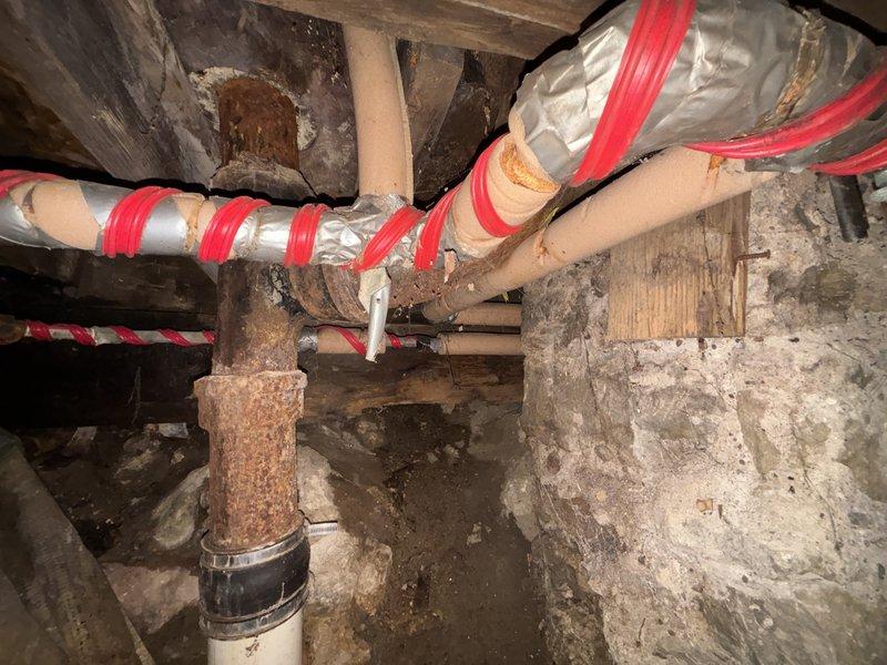 Inspected crawl space plumbing infrastructure and identified significant rust on metal pipe junctions and fittings. Observed moisture damage throughout area with visible mold/mildew on surrounding surfaces. Documented condition of water treatment system components including PVC piping and insulated lines, noting deterioration requiring remediation.