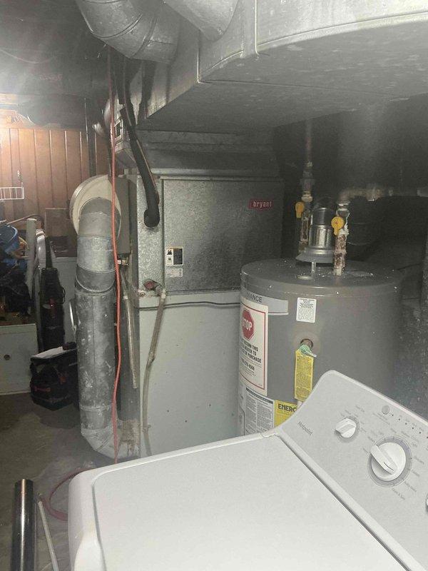 Conducted inspection of existing HVAC system in basement utility room, documenting aging furnace unit with connected ductwork and nearby water heater. Noted significant corrosion on plumbing components, particularly on metal pipes and fittings, with newer PVC sections having been previously installed. Recommended complete system replacement due to visible wear, rust damage, and potential efficiency concerns.